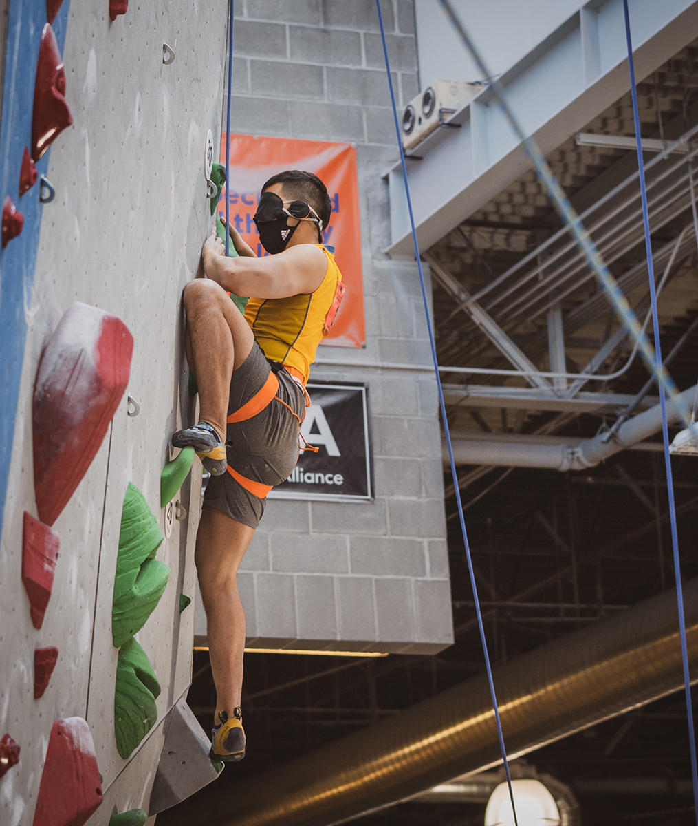 Paraclimbing athlete with a blindfold on is climbing up a indoor rock wall on top rope.
